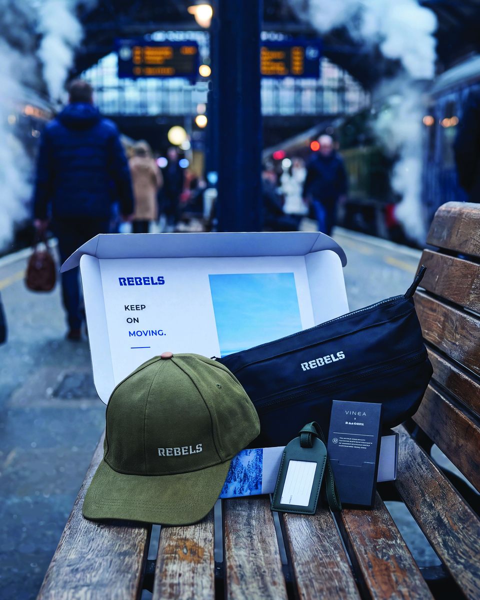 Geschenkset Winter on the move: Geschenkbox mit Kulturbeutel, Gepäckanhänger und Kappe auf einer Bank im Bahnhof